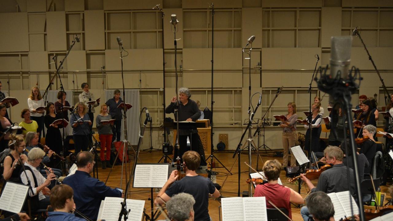 In einem Probenraum sind viele Mikrofone aufgebaut zwischen denen Musiker sitzen und etliche Sänger stehen und den Zeichen eines Dirigenten in der Mitte des Bildes folgen.