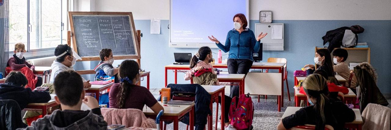 Schülerinnen und Schüler sitzen mit Masken und Abständen in einem Klassenraum. Vorne steht eine Lehrerin, ebenfalls mit Maske. Schülerinnen und Schüler sitzen mit Masken und Abständen in einem Klassenraum. Vorne steht eine Lehrerin, ebenfalls mit Maske.