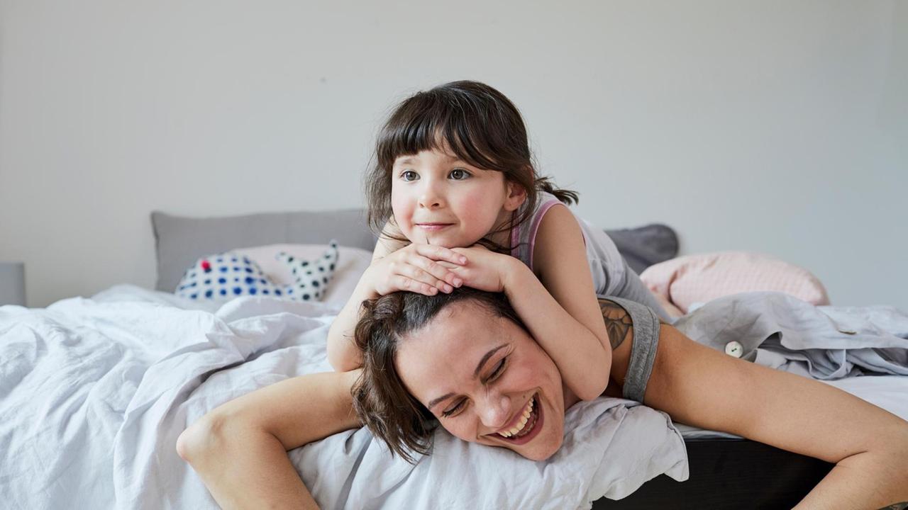 Mutter und Tochter liegen auf dem Bett, die Tochter liegt auf dem Rücken der lachenden Mutter. Mutter und Tochter liegen auf dem Bett, die Tochter liegt auf dem Rücken der lachenden Mutter.