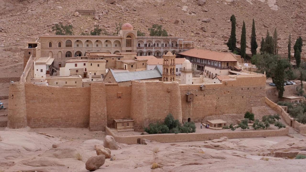 Rötliche Mauern fassen eine Klosteranlage in felsiger Wüstenlandschaft ein, im Innern: verschachtelte flache Häuser mit Ziegeldächern, einzelne Türme und ein Quergebäude mit Säulengängen. Rechts davon: Bäume, darunter einige schlanke hohe Zypressen. Rötliche Mauern fassen eine Klosteranlage in felsiger Wüstenlandschaft ein, im Innern: verschachtelte flache Häuser mit Ziegeldächern, einzelne Türme und ein Quergebäude mit Säulengängen. Rechts davon: Bäume, darunter einige schlanke hohe Zypressen.
