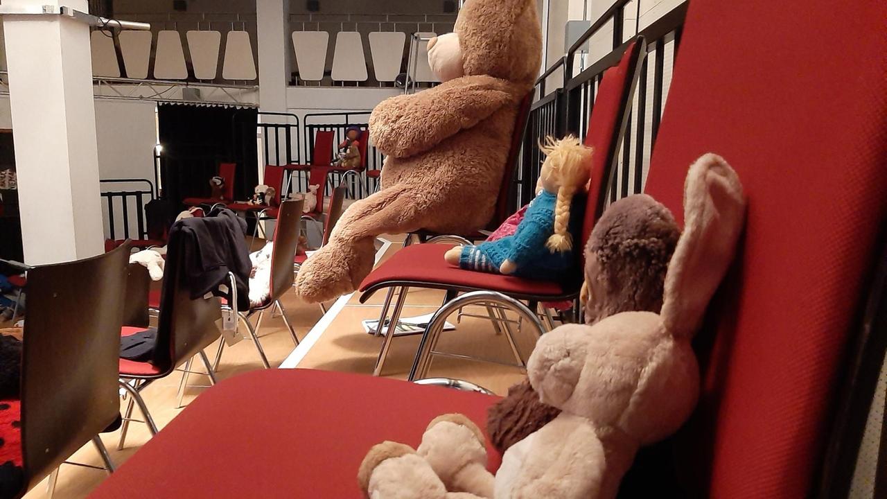 Auf den Publikumsplätzen (roten Stühlen) sitzen verschiende Kuscheltiere. Ein Plüschhase, ein menschengroßer Teddybär und eine Puppe.