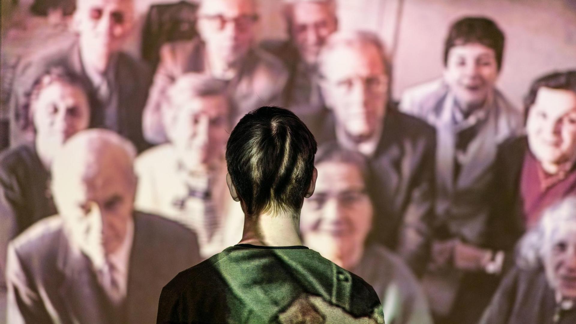 Ein junger Mann steht vor einer Video-Installation, auf der eine Menschengruppe zu sehen ist Ein junger Mann steht vor einer Video-Installation, auf der eine Menschengruppe zu sehen ist