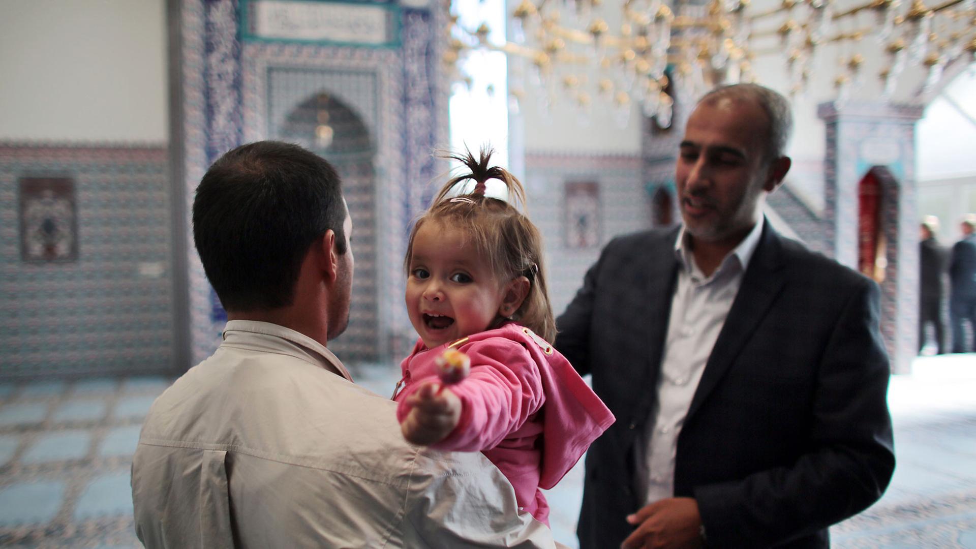 Ein syrischer Flüchtling spricht in einer Moschee mit einem anderen Mann. Er hält seine kleine Tochter auf dem Arm, die ihren Lutscher lachend in die Kamera hält.