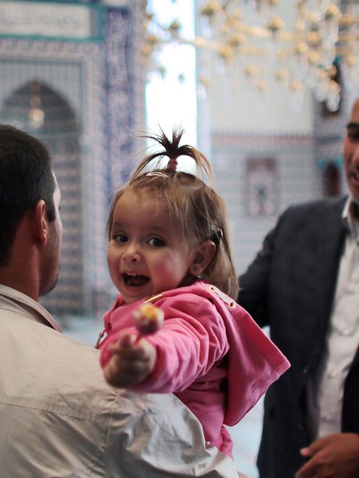 Ein syrischer Flüchtling spricht in einer Moschee mit einem anderen Mann. Er hält seine kleine Tochter auf dem Arm, die ihren Lutscher lachend in die Kamera hält.