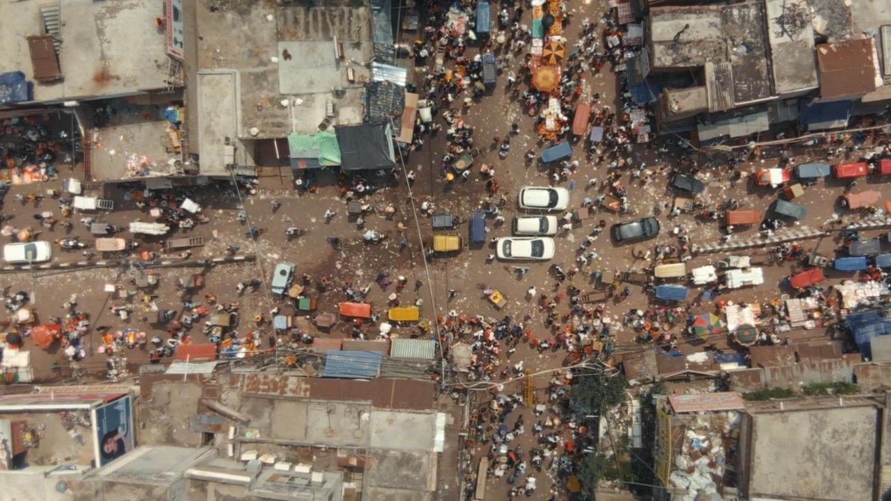 Ein Blick aus dem Luft nach unten zeigt eine wuselige Straßenszene in Indien. Mitten auf einer Kreuzung stehen drei weiße Autos. Fußgänger und andere motorisierte Fahrzeuge sind ebenso zu sehen wie die Dächer der anliegenden Häuser. Ein Blick aus dem Luft nach unten zeigt eine wuselige Straßenszene in Indien. Mitten auf einer Kreuzung stehen drei weiße Autos. Fußgänger und andere motorisierte Fahrzeuge sind ebenso zu sehen wie die Dächer der anliegenden Häuser.