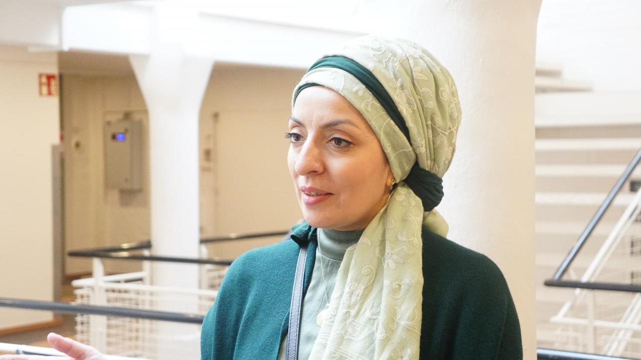 Eine Frau mit hellgrünem, besticktem Kopftuch blickt lächelnd, wie im Gespräch, an der Kamera vorbei. Hinter ihr erstreckt sich ein heller Raum. Eine Frau mit hellgrünem, besticktem Kopftuch blickt lächelnd, wie im Gespräch, an der Kamera vorbei. Hinter ihr erstreckt sich ein heller Raum.