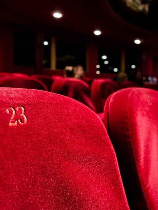 Das Foto zeigt in Großaufnahme leere rotsamtene Sitze in der Sitzreihe eines Theatersaals. Ein Sitz trägt die Nummer 23.