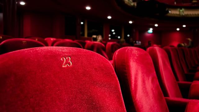 Das Foto zeigt in Großaufnahme leere rotsamtene Sitze in der Sitzreihe eines Theatersaal. Ein Sitz trägt die Nummer 23.