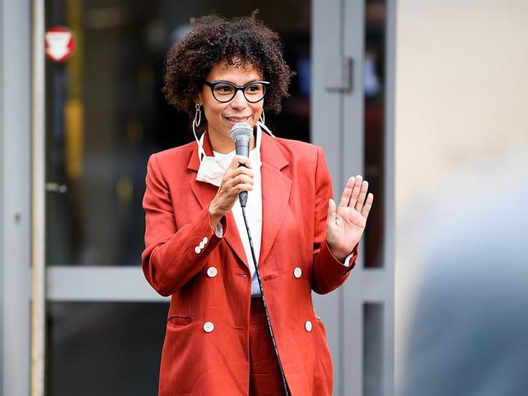 Julia Wissert hebt grüßend die Hand und lächelt das Publikum an. Sie trägt einen roten Blazer, eine markante schwarze Brille und die Haare offen. Julia Wissert hebt grüßend die Hand und lächelt das Publikum an. Sie trägt einen roten Blazer, eine markante schwarze Brille und die Haare offen.