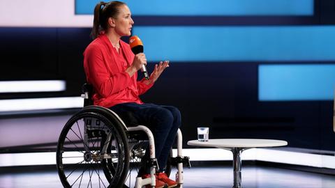 Eine junge Frau ist zu Gast in einem TV-Studio, sitzt in einem Rollstuhl und spricht in ein Mikrofon.