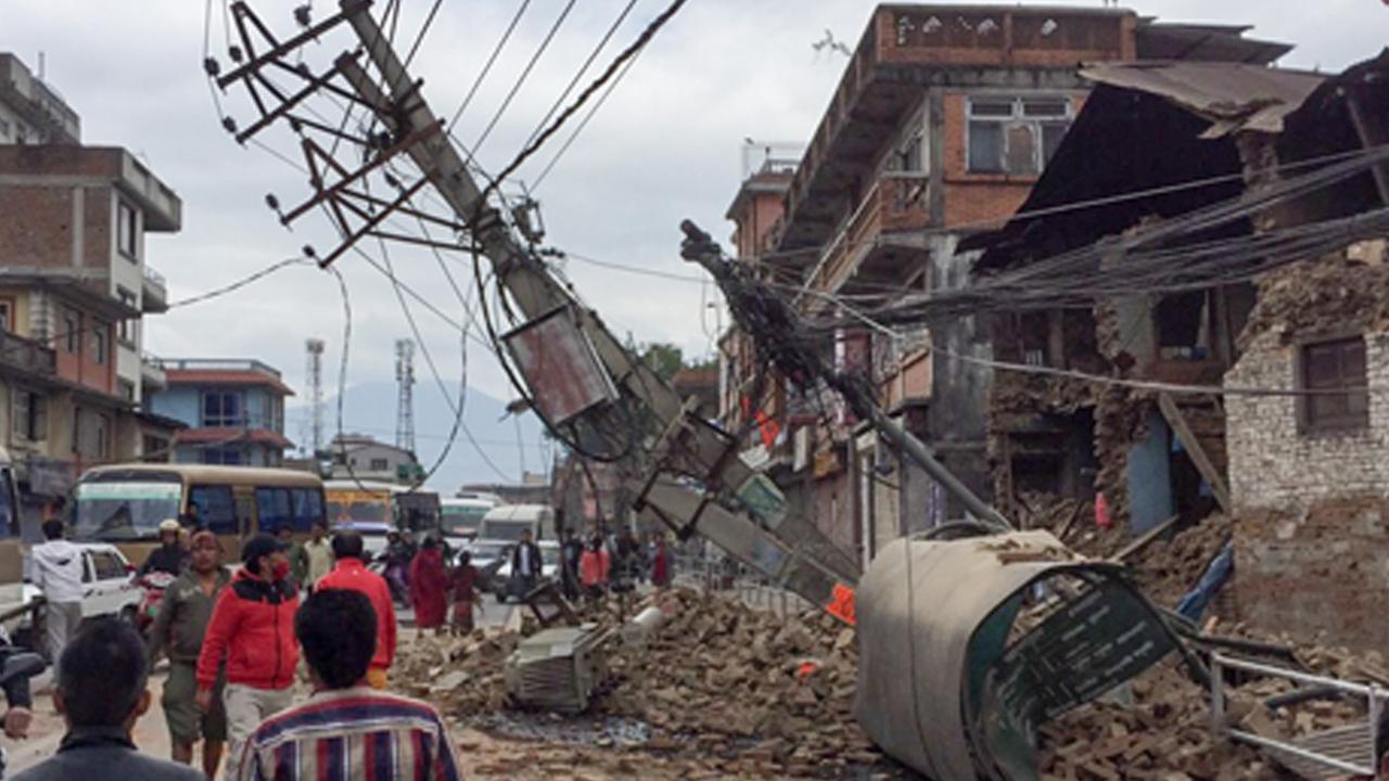 Eingestürzte Häuser in Kathmandu. Passanten laufen vorbei. Auch ein Strommast ist umgestürzt.
