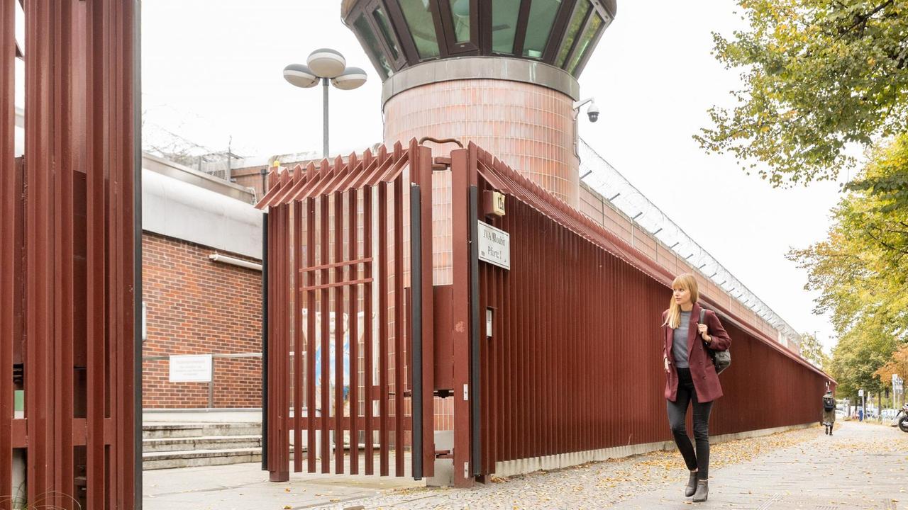Eine junge Frau mit langen blonden Haaren und weinrotem Blazer geht an einer Gefängnismauer entlang auf ein großes offenes Tor zu. Eine junge Frau mit langen blonden Haaren und weinrotem Blazer geht an einer Gefängnismauer entlang auf ein großes offenes Tor zu.