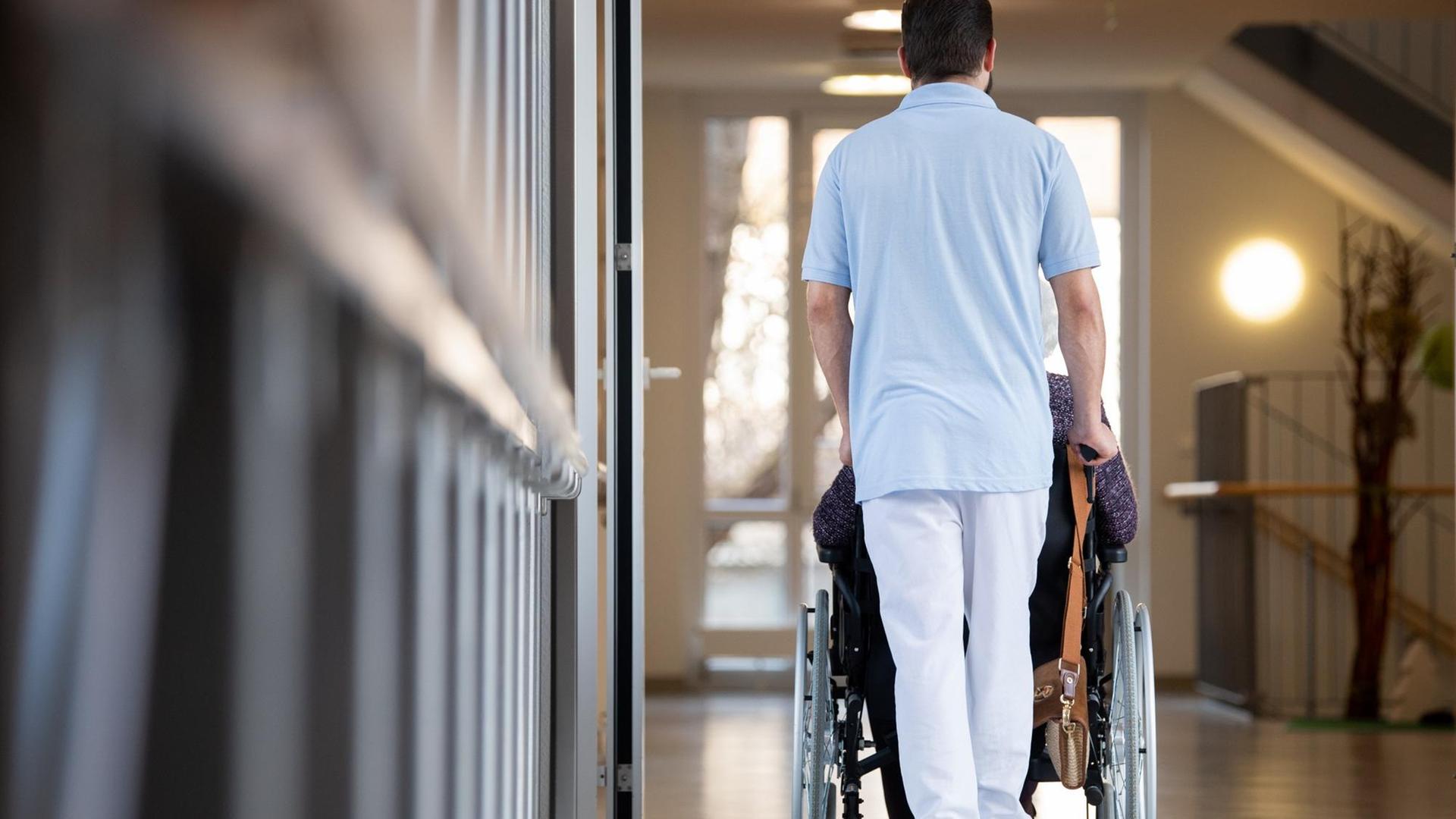 Ein Pfleger schiebt eine Bewohnerin eines Pflegeheims in einem Rollstuhl über den Flur. Ein Pfleger schiebt eine Bewohnerin eines Pflegeheims in einem Rollstuhl über den Flur.