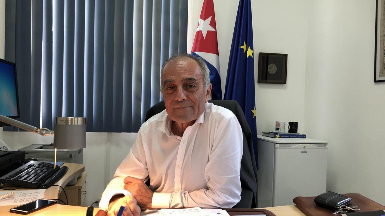Ein älterer Herr im weißen Hemd sitzt in seinem Büro am Schreibtisch. Hinter ihm stehen die EU- und die kubanische Flagge.