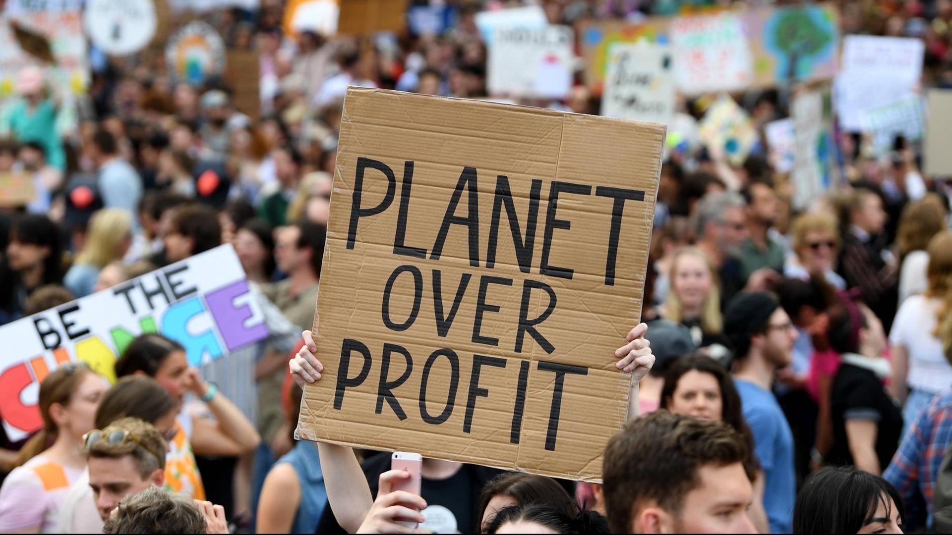 Bei den Protesten des Klimastreiks ist ein Schild zu sehen auf dem "Planet over Profits" zu lesen ist. Dahinter ist eine große Menschenmenge zu erkennen.