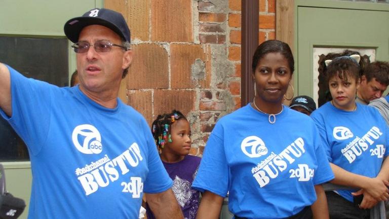 John George steht gemeinsam mit zwei Frauen - alle in knallblauen T-Shirts auf denen "Bustup" steht - bei einer öffentlichen Aktion in einer Reihe. John George steht gemeinsam mit zwei Frauen - alle in knallblauen T-Shirts auf denen "Bustup" steht - bei einer öffentlichen Aktion in einer Reihe.