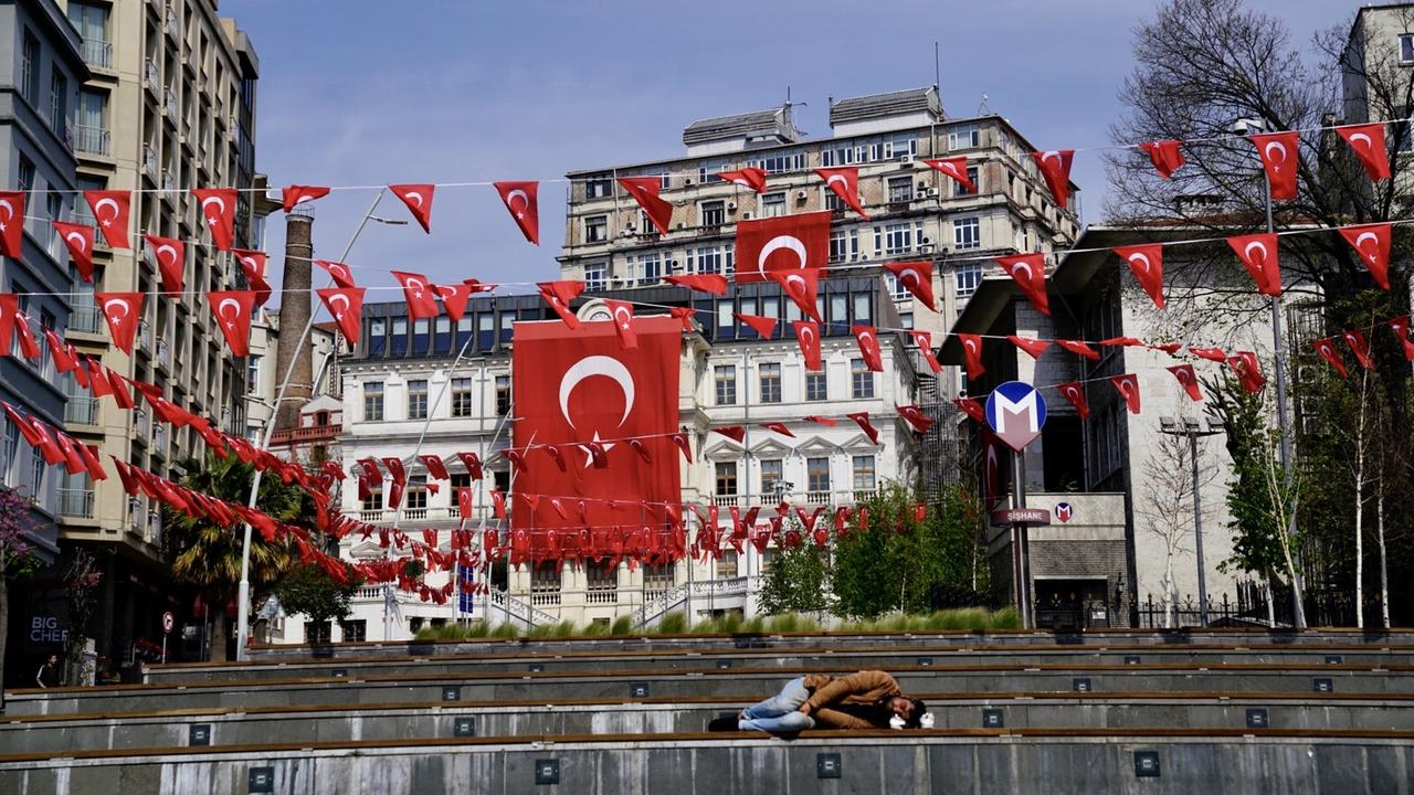 Zahlreiche leere Bänke unter vielen rot-weißen kleinen türkischen Flaggen in Istanbul. Auf einer der Bänke ganz vorne schläft ein Obdachloser. Zahlreiche leere Bänke unter vielen rot-weißen kleinen türkischen Flaggen in Istanbul. Auf einer der Bänke ganz vorne schläft ein Obdachloser.