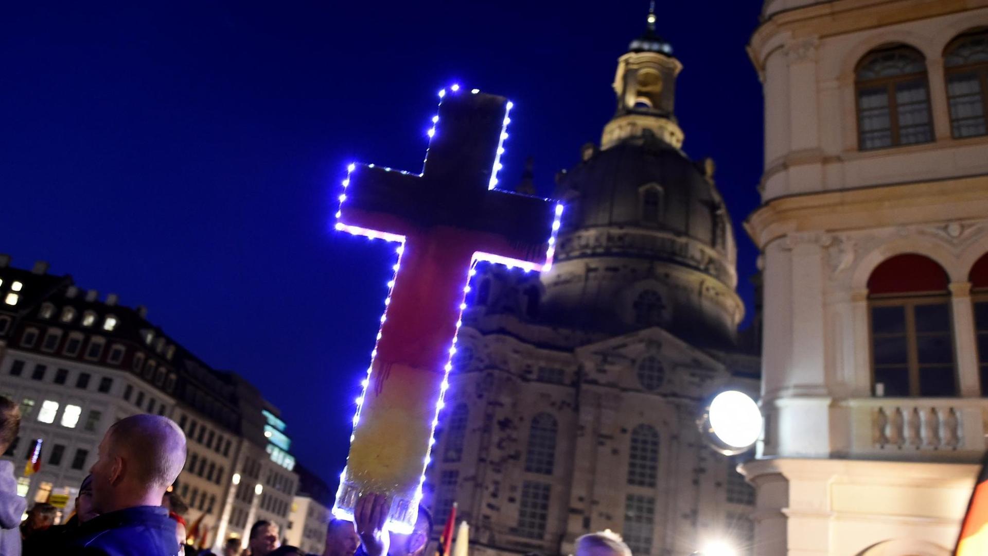 Ein Anhänger der PEGIDA-Bewegung hält auf der Protestkundgebung vor der Frauenkirche in Dresden ein in deutschen Nationalfarben gemaltes Kreuz in der Hand. Ein Anhänger der PEGIDA-Bewegung hält auf der Protestkundgebung vor der Frauenkirche in Dresden ein in deutschen Nationalfarben gemaltes Kreuz in der Hand.