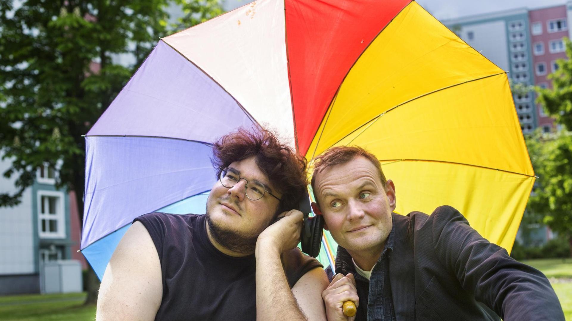 Devid Striesow und Axel Ranisch lehnen sich unter einem bunten Regenschirm aneinander und lauschen den Tönen aus einem Kopfhörer. Devid Striesow und Axel Ranisch lehnen sich unter einem bunten Regenschirm aneinander und lauschen den Tönen aus einem Kopfhörer.