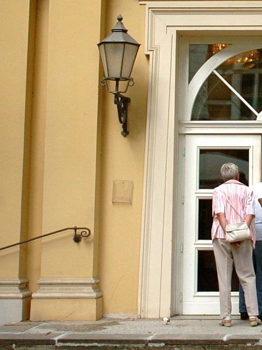 Zwei Theaterbesucher blicken neugierig durch die Eingangstür der Alten Oper in Erfurt.