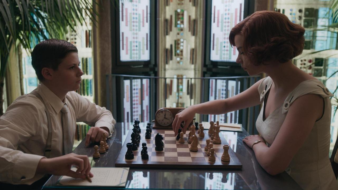 Anya Taylor-Joy, in der Rolle der Elizabeth "Beth" Harmon, sitzte am Tisch und spielt gegen einen jungen Mann Schach. Anya Taylor-Joy, in der Rolle der Elizabeth "Beth" Harmon, sitzte am Tisch und spielt gegen einen jungen Mann Schach.