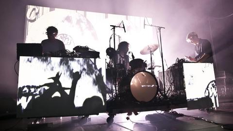 Eine Konzertbühne: In der Mitte ein Schlagzeuger, gerahmt von zwei Musikern an Mischpulten. Das Licht beleuchtet die Szene in einer Weise, dass sie wie das Negativ einer Schwarzweißfotografie erscheint.