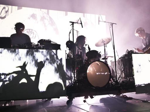 Eine Konzertbühne: In der Mitte ein Schlagzeuger, gerahmt von zwei Musikern an Mischpulten. Das Licht beleuchtet die Szene in einer Weise, dass sie wie das Negativ einer Schwarzweißfotografie erscheint.
