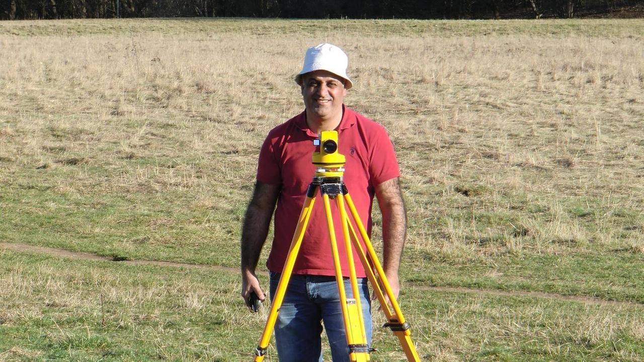 Ismael Ibraheem bei der Feldarbeit. Er steht auf einer Wiese vor sich seine Messapparatur und lacht in die Kamera. Die Sonne scheint, er trägt einen kleinen Sonnenhut. Ismael Ibraheem bei der Feldarbeit. Er steht auf einer Wiese vor sich seine Messapparatur und lacht in die Kamera. Die Sonne scheint, er trägt einen kleinen Sonnenhut.