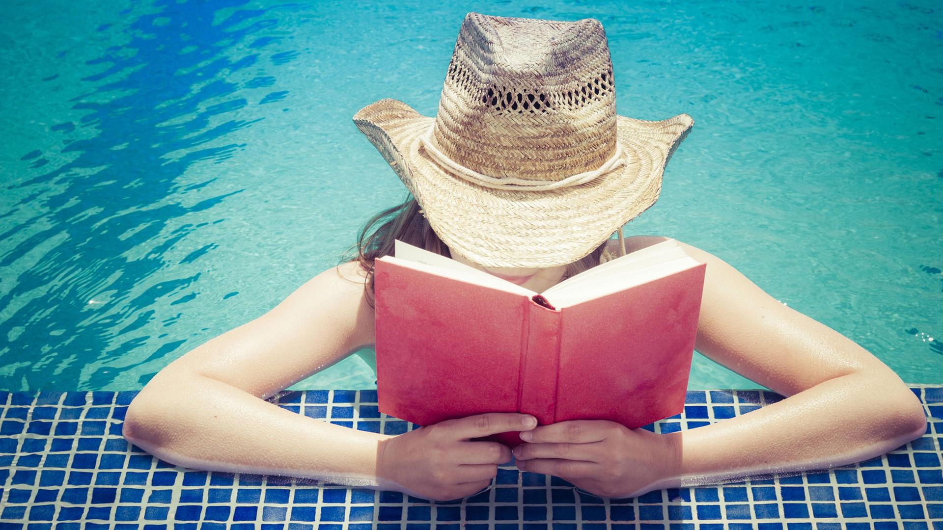 Eine Frau mit Strohhut liest am Beckenrand eines Schwimmbeckens ein Buch. Eine Frau mit Strohhut liest am Beckenrand eines Schwimmbeckens ein Buch.
