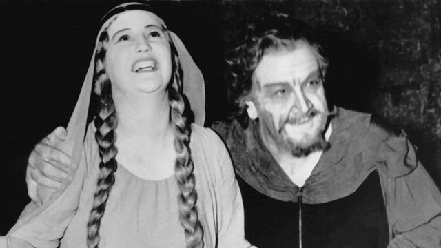 Eine Frau (links) mit langen Zöpfen und im langen Kleid steht neben bärtigem Mann auf einer Opernbühne.