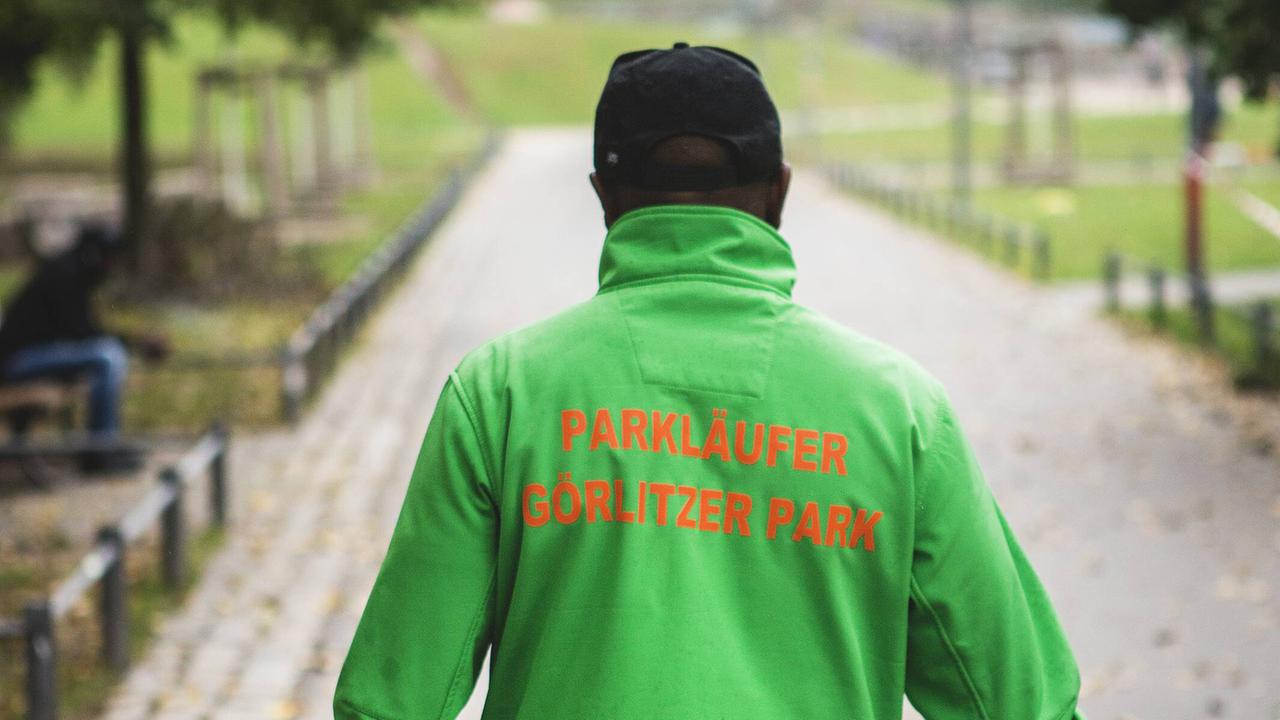 Mann sieht einen Mann von hinten in gründer Jacke, auf dem "Parkläufer" steht. Parkläufer arbeiten im Berliner Görlitzer Park als eine Art Sozialvermittler zwischen Drogendealern und Polizei. Mann sieht einen Mann von hinten in gründer Jacke, auf dem "Parkläufer" steht. Parkläufer arbeiten im Berliner Görlitzer Park als eine Art Sozialvermittler zwischen Drogendealern und Polizei.