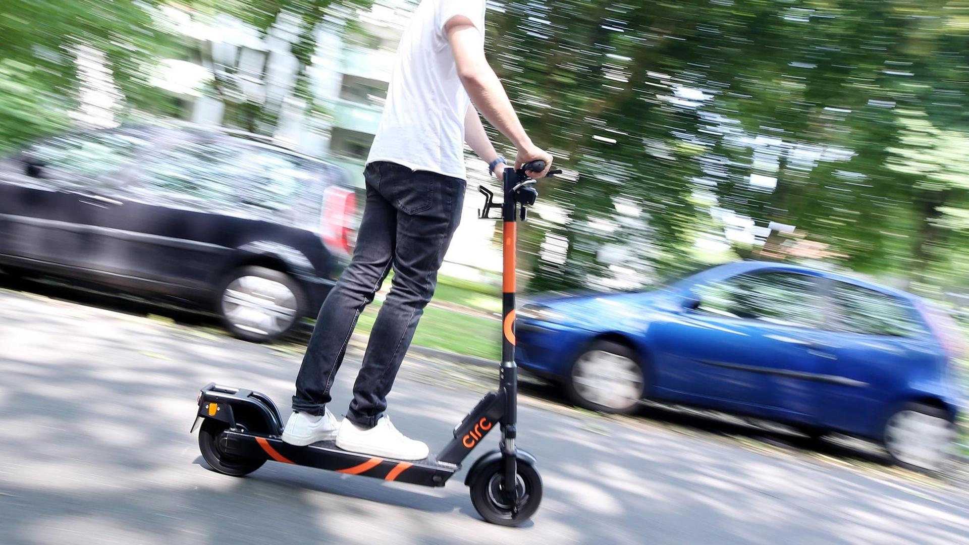Ein Mann macht eine Probefahrt mit einem blauen E-Scooter, einem Elektroroller. Im Hintergrund sind parkende Autos zu sehen. Ein Mann macht eine Probefahrt mit einem blauen E-Scooter, einem Elektroroller. Im Hintergrund sind parkende Autos zu sehen.