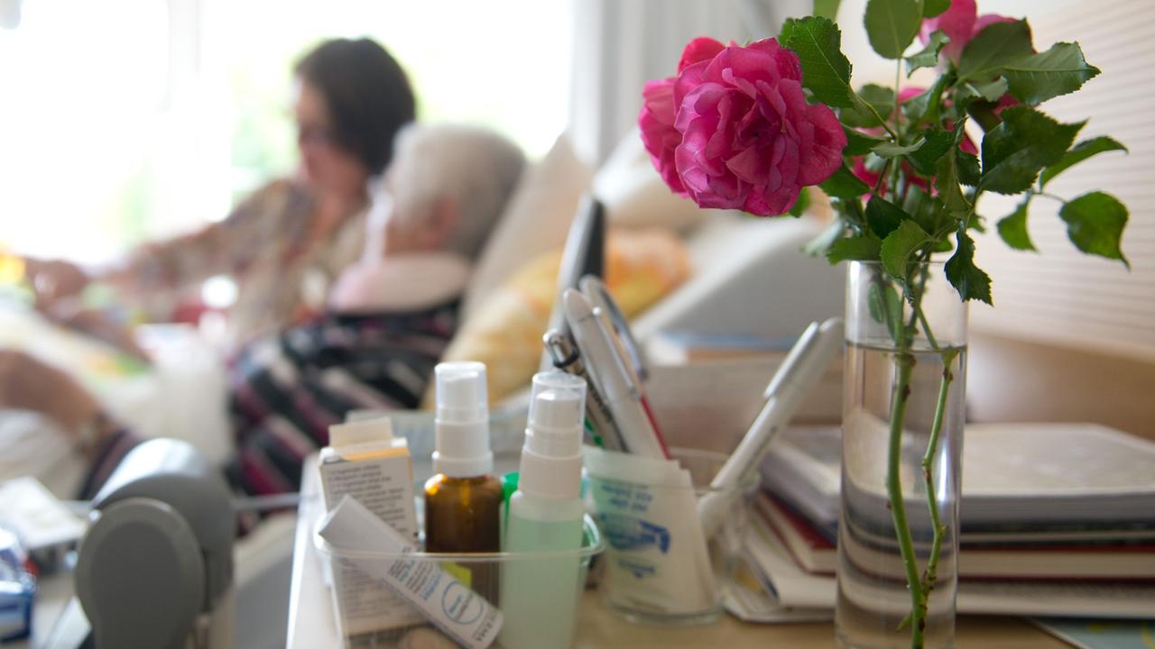 Im Vordergrund eine Rose, im Hintergrund ein Krankenbett mit einer alten Frau und einer jüngeren am Bett. Im Vordergrund eine Rose, im Hintergrund ein Krankenbett mit einer alten Frau und einer jüngeren am Bett.