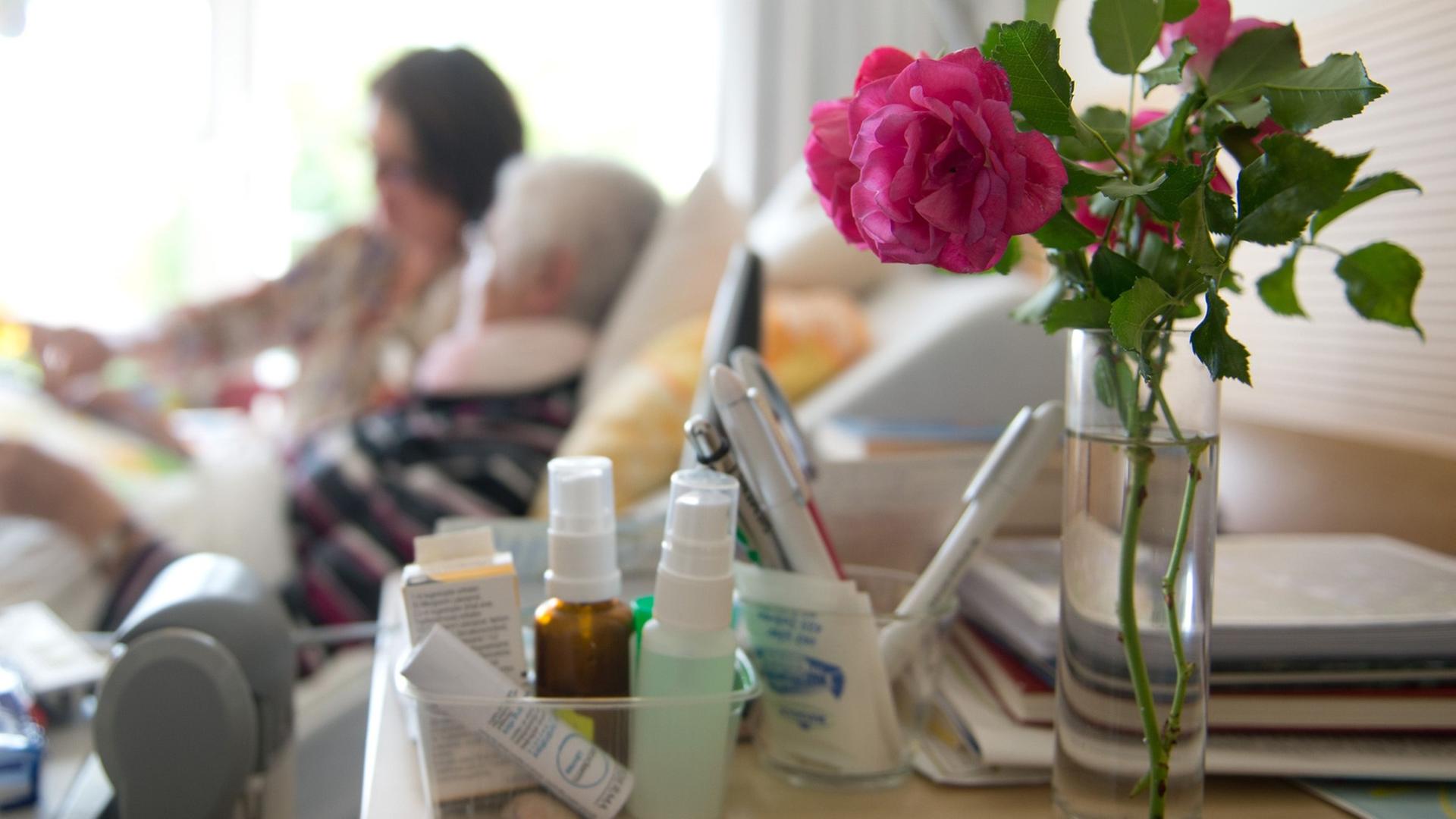 Im Vordergrund eine Rose, im Hintergrund ein Krankenbett mit einer alten Frau und einer jüngeren am Bett. Im Vordergrund eine Rose, im Hintergrund ein Krankenbett mit einer alten Frau und einer jüngeren am Bett.