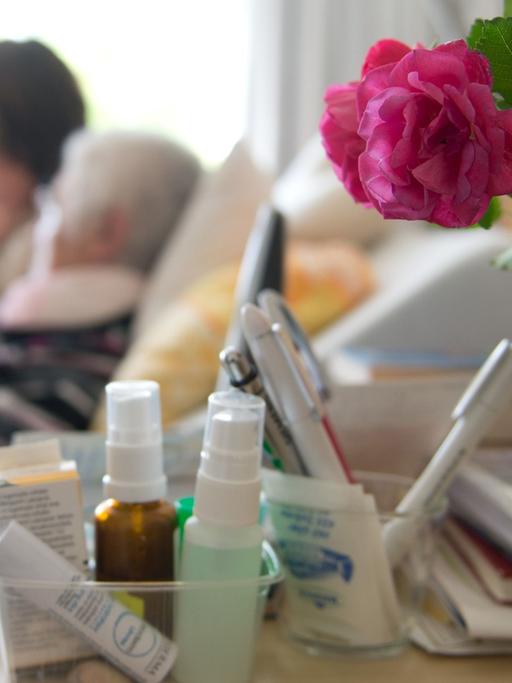 Im Vordergrund eine Rose, im Hintergrund ein Krankenbett mit einer alten Frau und einer jüngeren am Bett.