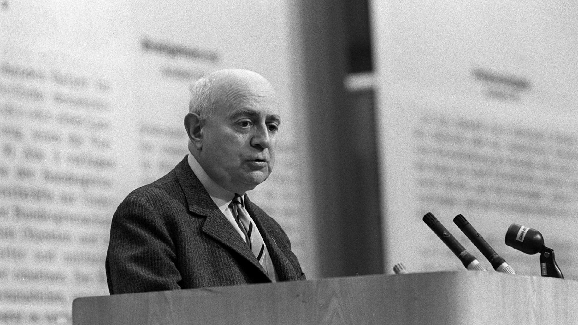 Der Philosoph Theodor W. Adorno mit nachdenklichem Blick vor einer Reihe Mikrofone, an der Rückwand des großen Saales erkennt man verschwommene Schrift. Der Philosoph Theodor W. Adorno mit nachdenklichem Blick vor einer Reihe Mikrofone, an der Rückwand des großen Saales erkennt man verschwommene Schrift.