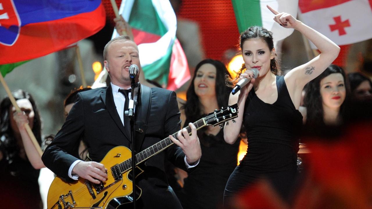 Stefan Raab singt im Anzug mit Gitarre in ein Mikrofon, neben ihm singt Lena im schwarzen Kleid. Um sie herum stehen Menschen, einige tragen Fahnen von Teilnehmerländern des Eurovission Song Contest. Stefan Raab singt im Anzug mit Gitarre in ein Mikrofon, neben ihm singt Lena im schwarzen Kleid. Um sie herum stehen Menschen, einige tragen Fahnen von Teilnehmerländern des Eurovission Song Contest.