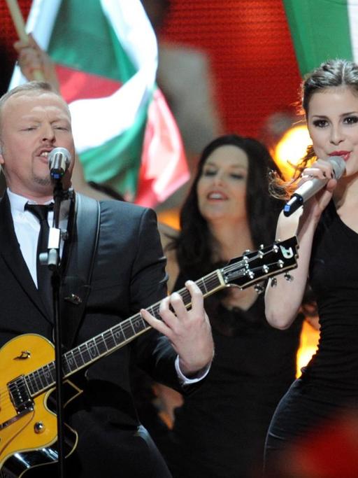 Stefan Raab singt im Anzug mit Gitarre in ein Mikrofon, neben ihm singt Lena im schwarzen Kleid. Um sie herum stehen Menschen, einige tragen Fahnen von Teilnehmerländern des Eurovission Song Contest.
