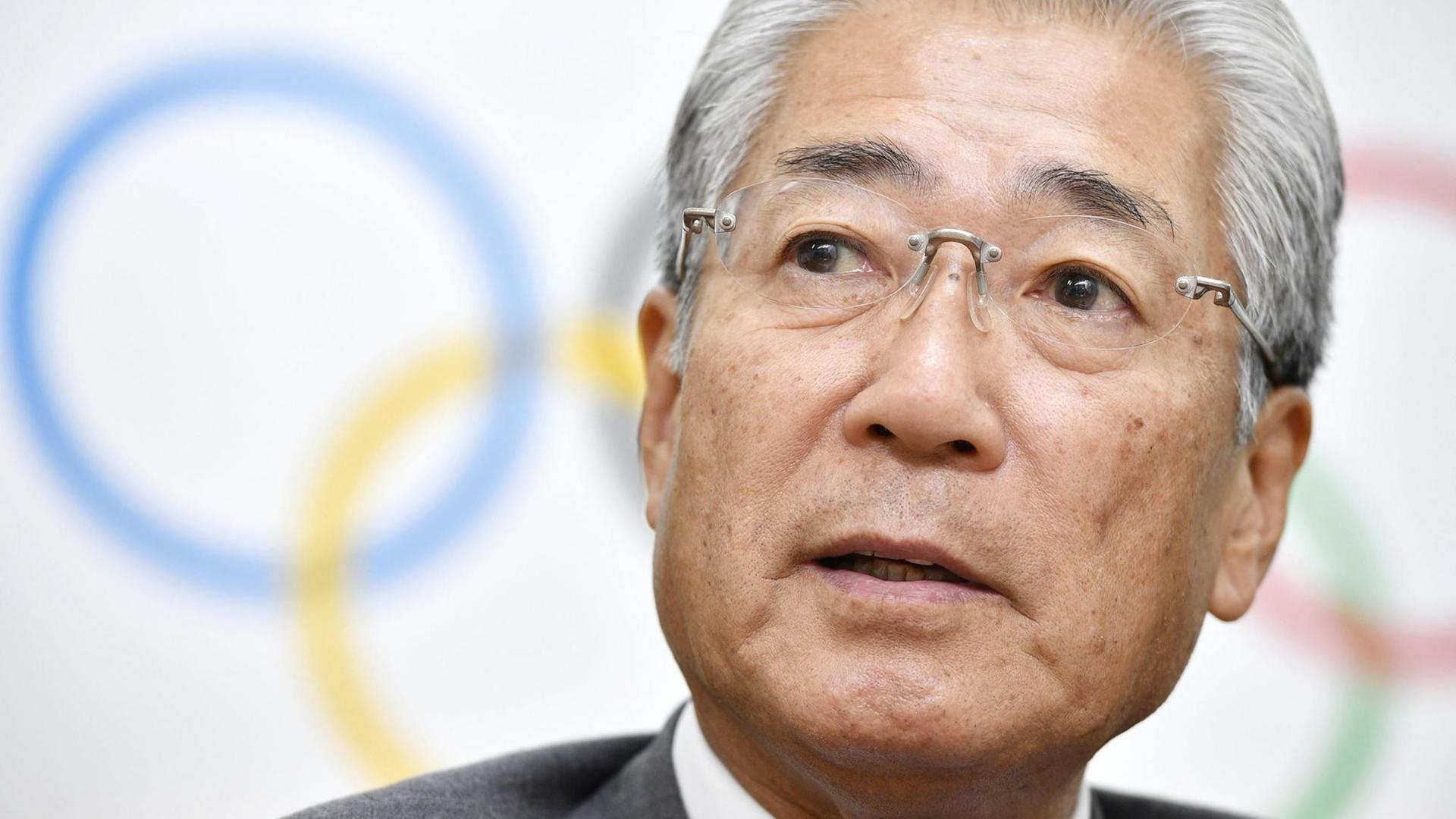 Der Präsident des japanischen Olympia-Komitees Takeda im Porträt, dahinter die Olympischen Ringe.