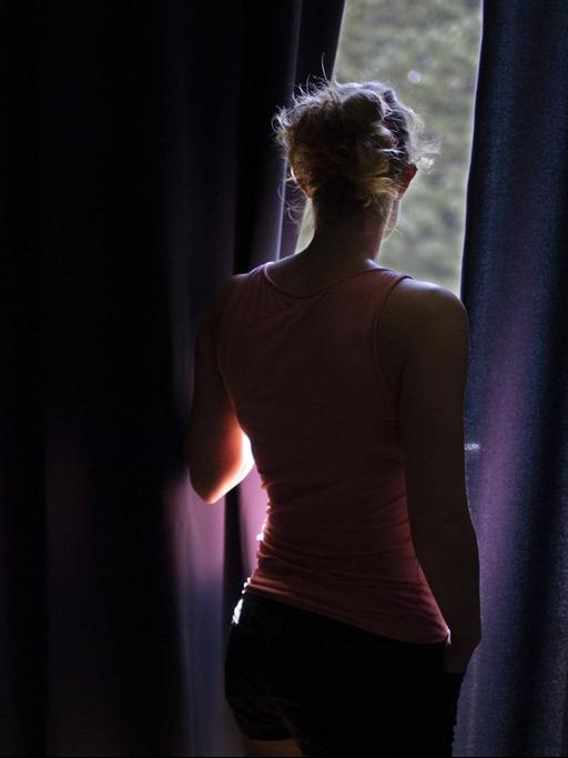 Eine Frau schaut zwischen dicken Vorhängen zum Fenster heraus.