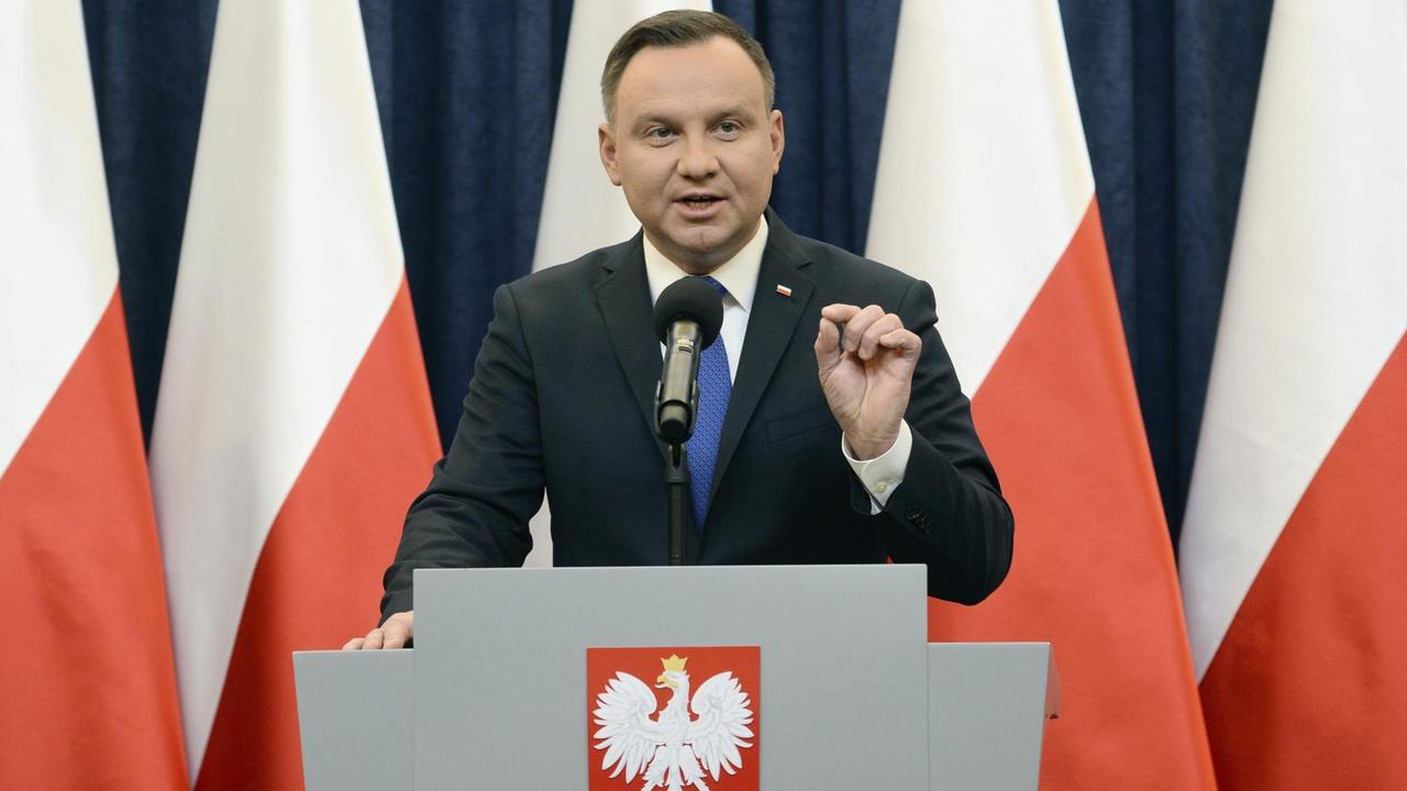 Das Bild zeigt Polens Präsidenten Andrzej Duda, er steht vor polnischen Fahnen in weiß-rot an einem Rednerpult.