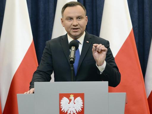 Das Bild zeigt Polens Präsidenten Andrzej Duda, er steht vor polnischen Fahnen in weiß-rot an einem Rednerpult.