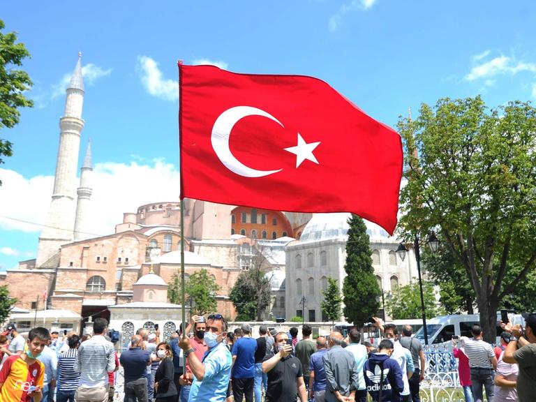 Blick auf die Hagia Sophia in Istanbul. Im Vordergrund weht eine türkische Flagge, die ein Mann auf dem Platz in die Höhe hält.
