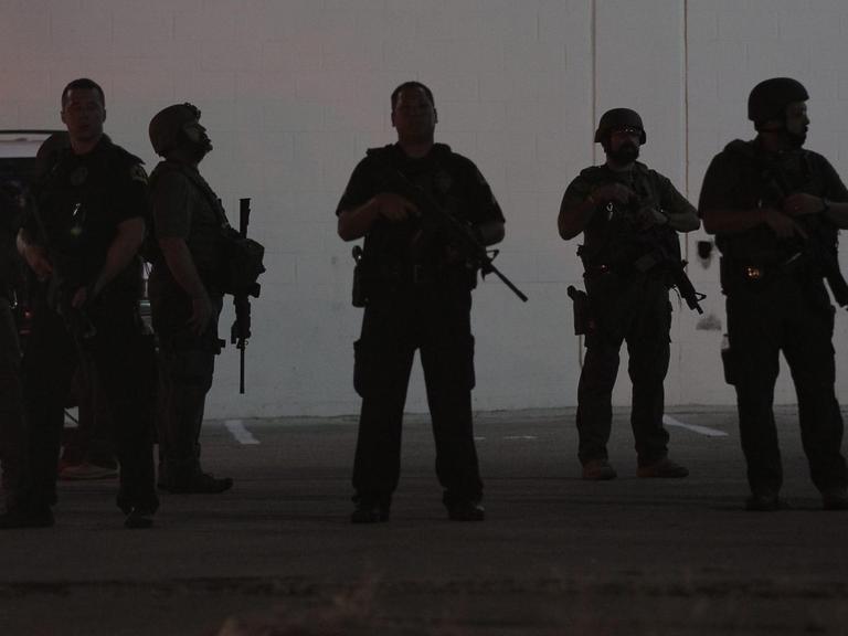 Silhouetten von schwer bewaffneten Polizisten vor einer grauen Wand.