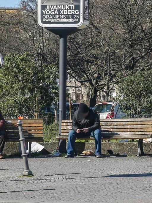 Drei Obdachlose sitzen in Berlin mit großem Abstand auf Parkbänken auf einem Platz in Kreuzberg.