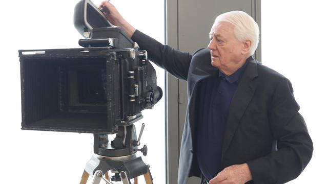 Alexander Kluge steht neben seiner Filmkamera und erklärt ihre Funktionsweise.