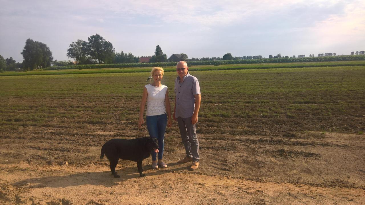 Das Ehepar Romaniuk steht auf einem Feld mit Hund und lacht. Das Ehepar Romaniuk steht auf einem Feld mit Hund und lacht.