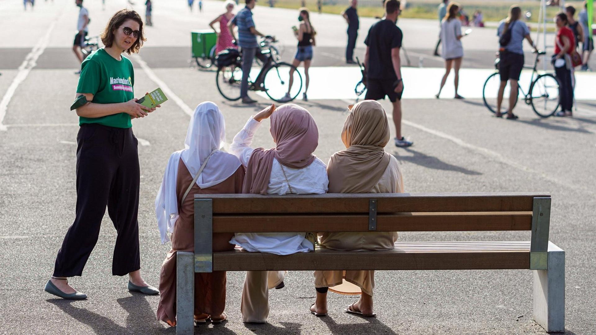 Eine Anhängerin der Grünen geht auf drei Frauen mit Kopftuch zu, die auf einer Parkbank auf dem Tempelhofer Feld sitzen