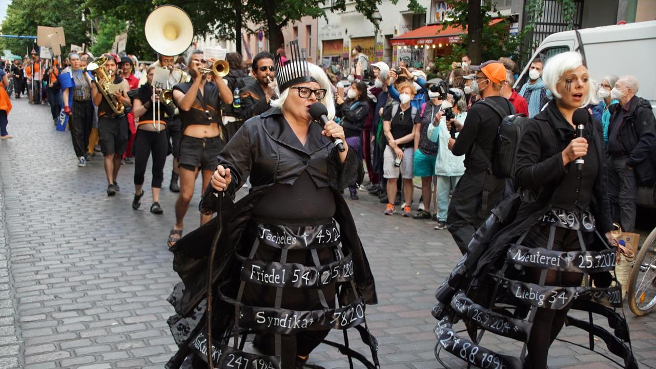 Schwarz gekleidete Schauspieler:innen gehen eine Straße entlang und singen. Schwarz gekleidete Schauspieler:innen gehen eine Straße entlang und singen.