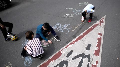 Kinder malen mit Kreide auf einer Straße, auf der sonst rund um die Uhr Autos fahren, ein Kind Kinder malen mit Kreide auf einer Straße, auf der sonst rund um die Uhr Autos fahren.hält einen Ball unter seinem Fuß.